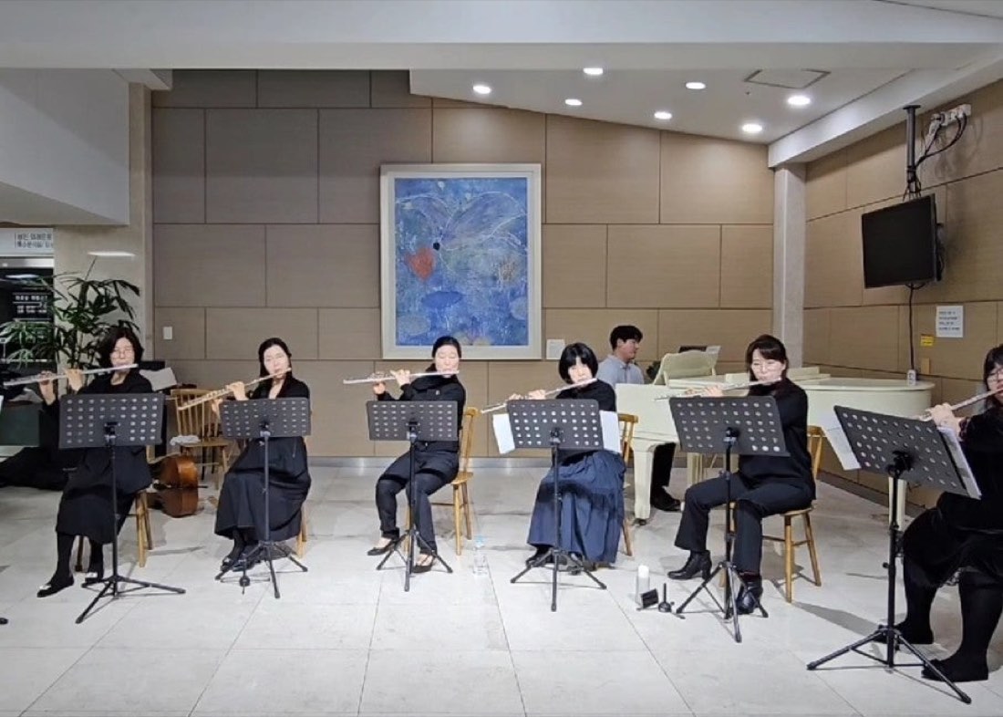 병원 로비에서 검은 옷을 입은 플루트 연주자 여섯 명이 보면대 앞에 앉아 연주하는 모습